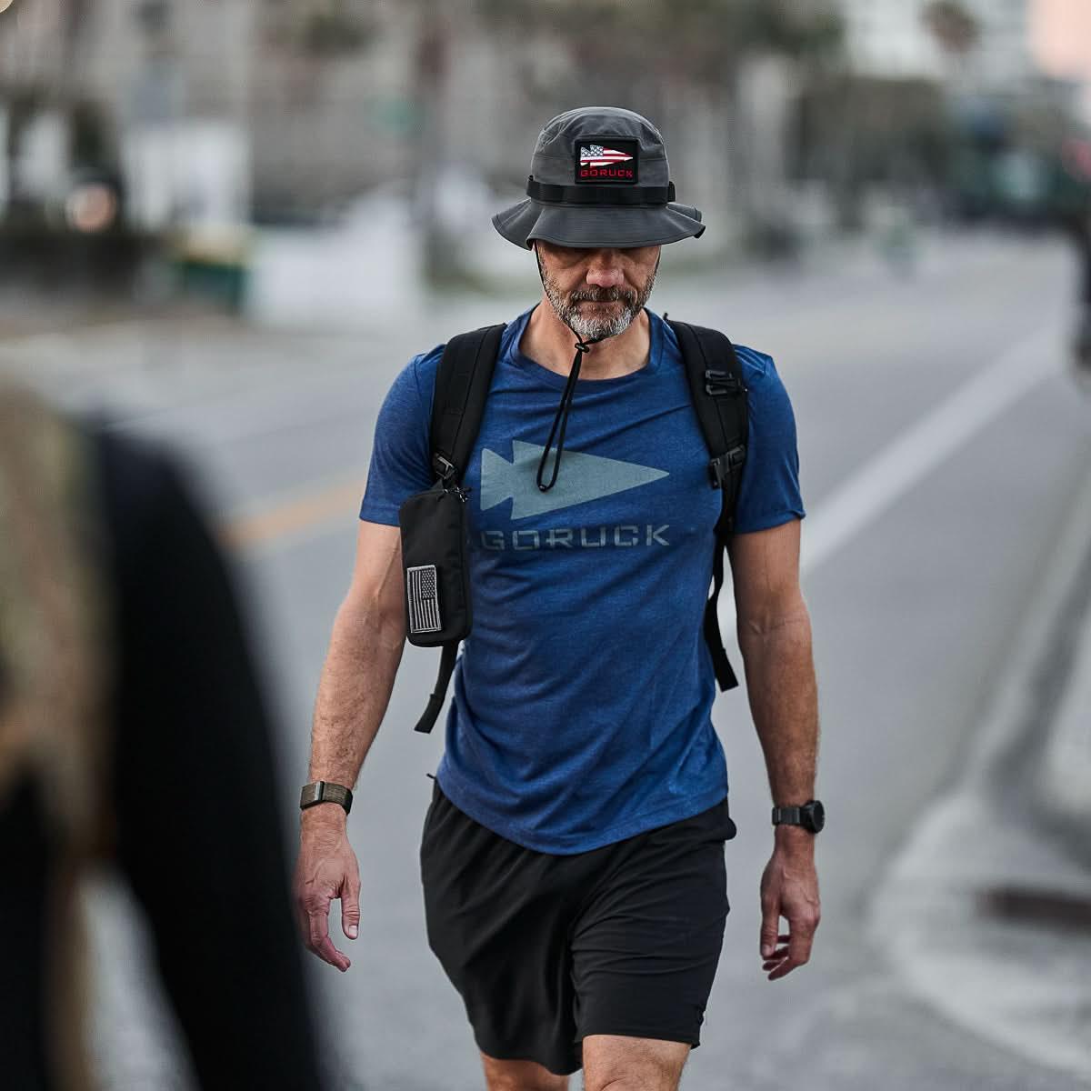 Man walking outdoors wearing GORUCK navy blue t-shirt, black bucket hat with American flag patch, and carrying backpack