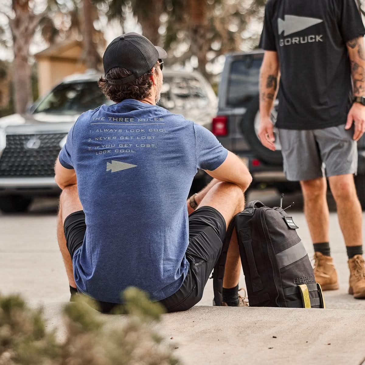 Man wearing GORUCK navy 'The Three Rules' t-shirt sitting outdoors with black GORUCK backpack in urban setting