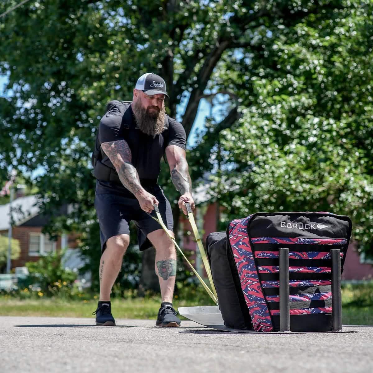 Bearded man with tattoos pulling a GORUCK weighted sled outdoors on pavement, surrounded by trees and houses