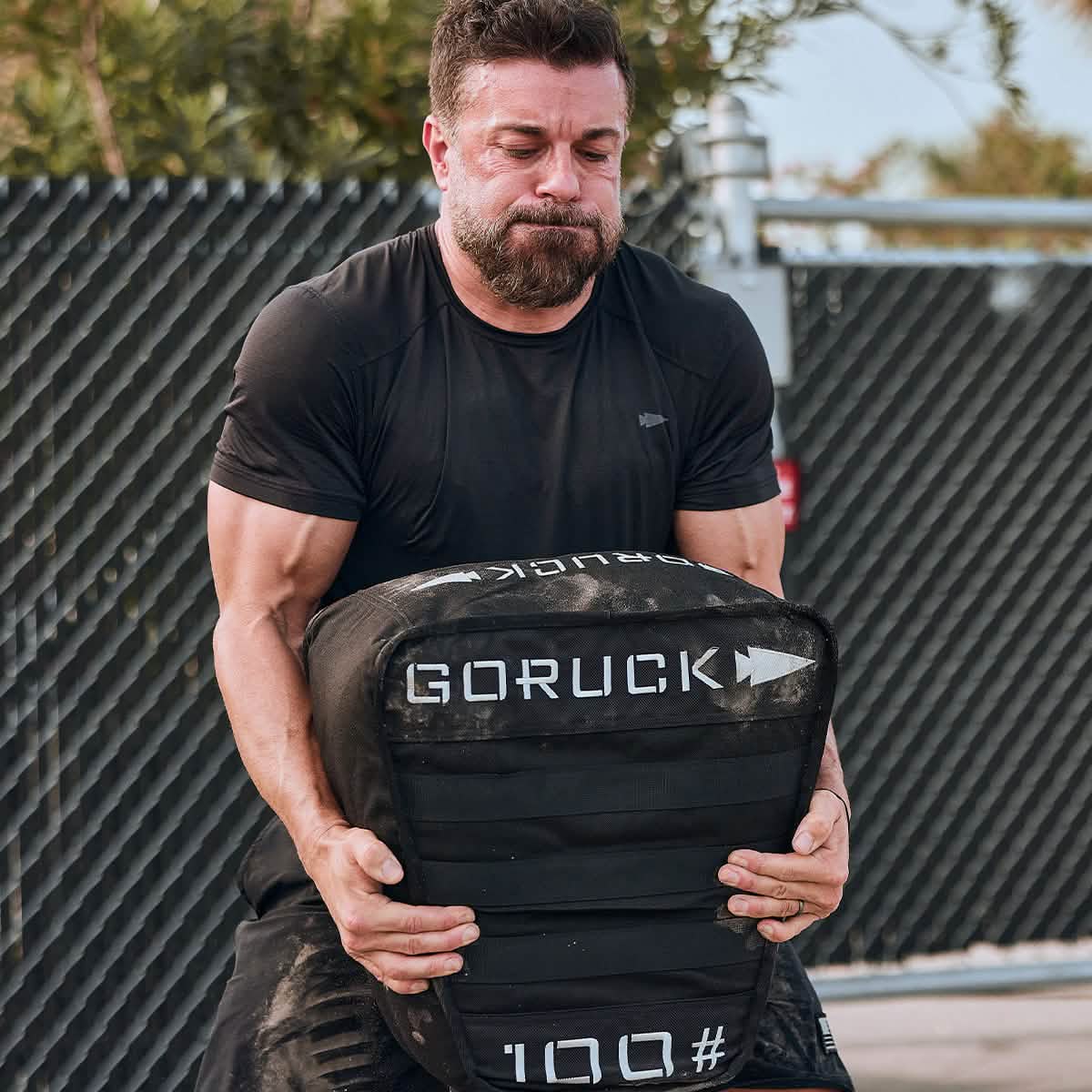 A man lifts a heavy 100-pound GORUCK sandbag outdoors, straining in his Men’s USA Performance Tee - ToughMesh, a featherweight shirt made from quick drying performance fabric.