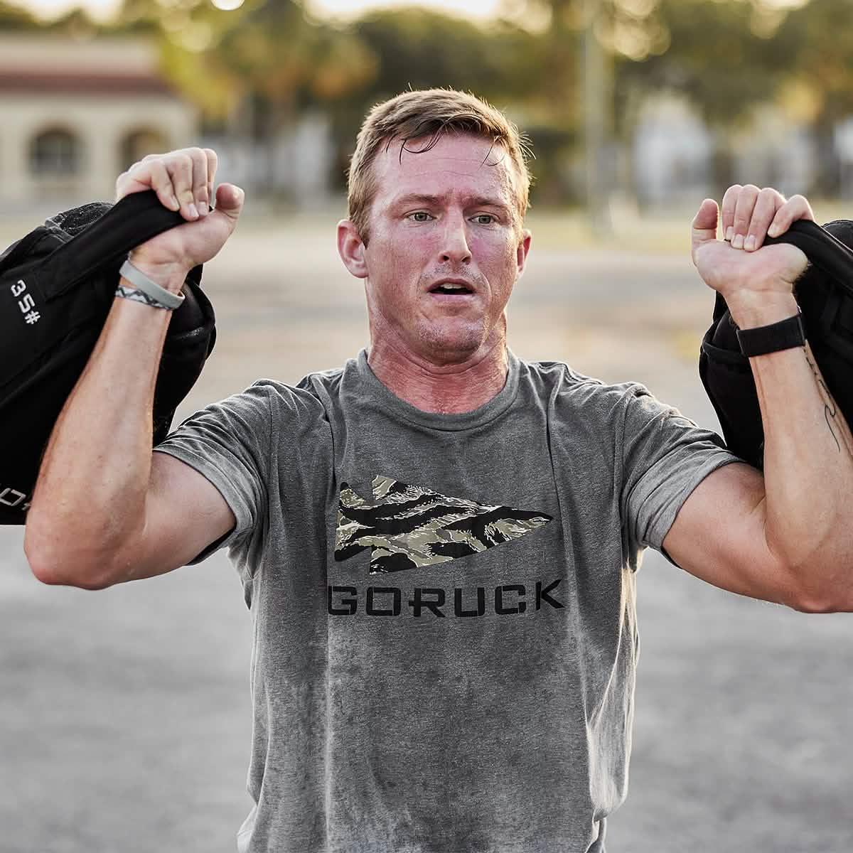 Man wearing GORUCK shirt outdoors, lifting weighted sandbags during rucking workout