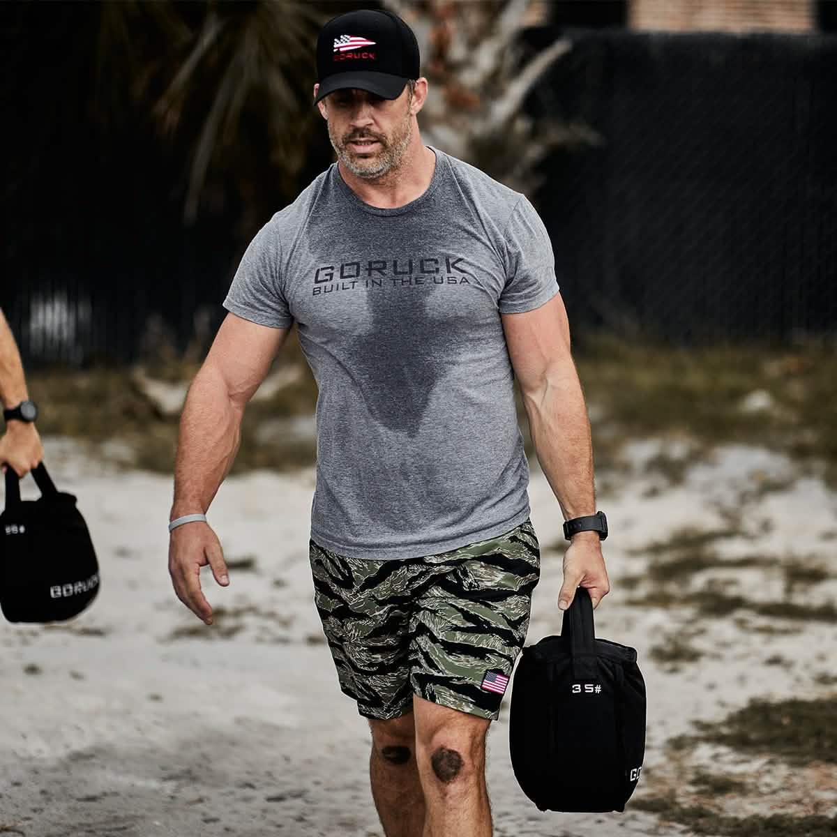 Man wearing GORUCK shirt and hat carrying weighted ruck bag outdoors on sandy terrain