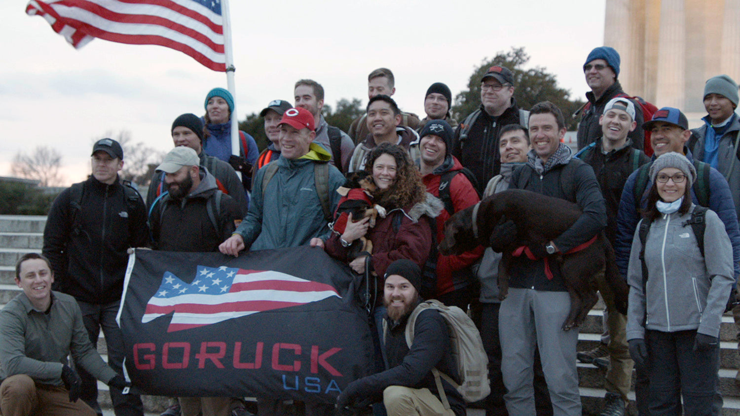 Group of people outdoors posing with US flag and GORUCK USA banner, some smiling, two holding dogs.