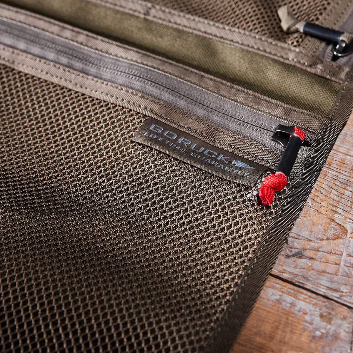 A close-up of the GORUCK Wire Dopp, a mesh pouch with a zipper, resting on a wooden surface. Crafted from durable CORDURA, this pouch is perfect for organized carry. The zipper pull has a red cord for easy opening, making it an ideal travel essential.