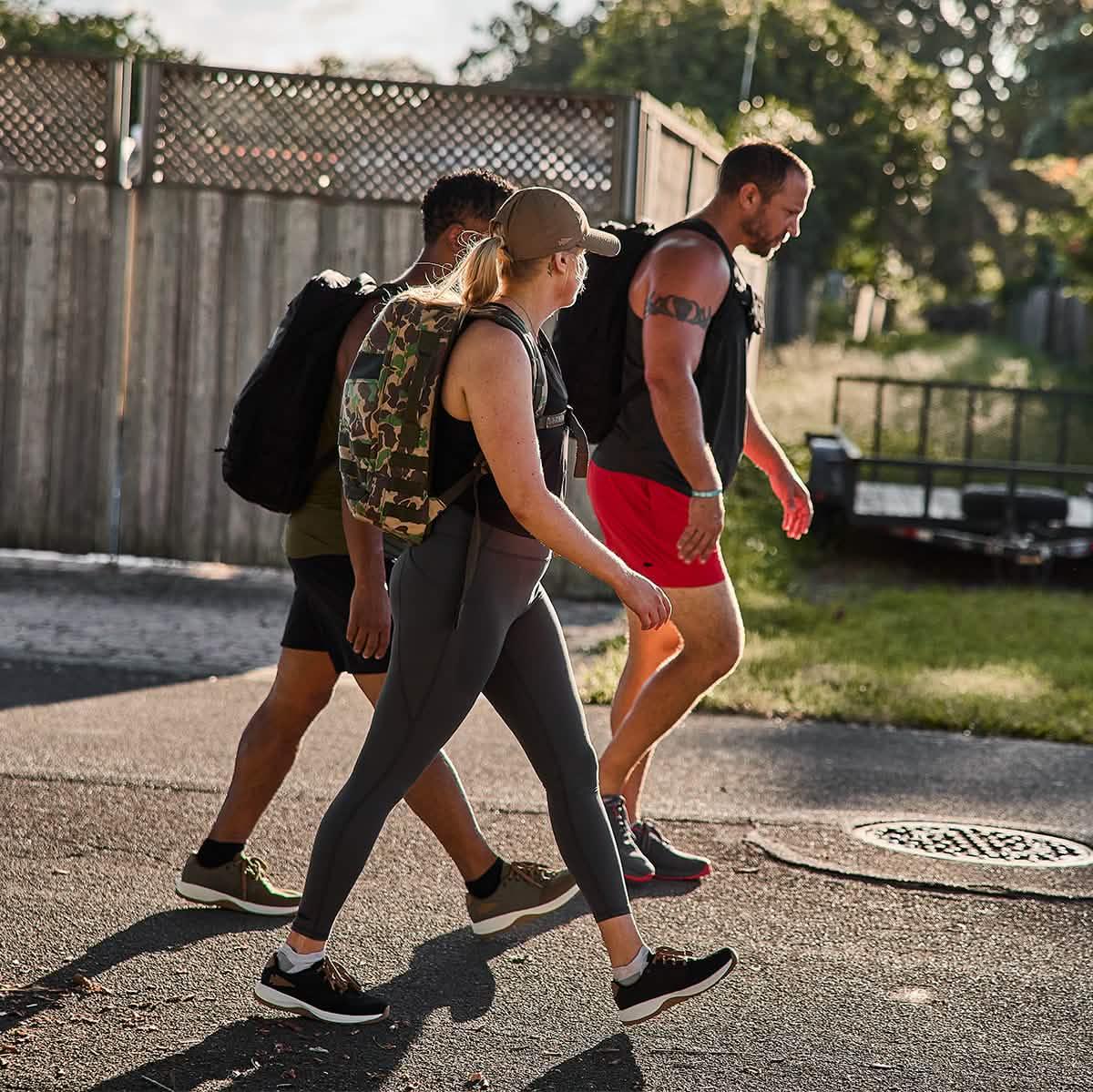 Three people walking outdoors wearing GORUCK weighted backpacks and athletic clothing during golden hour