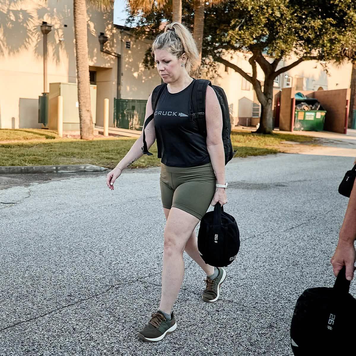 Woman rucking outdoors with GORUCK gear, wearing a black tank top and olive shorts
