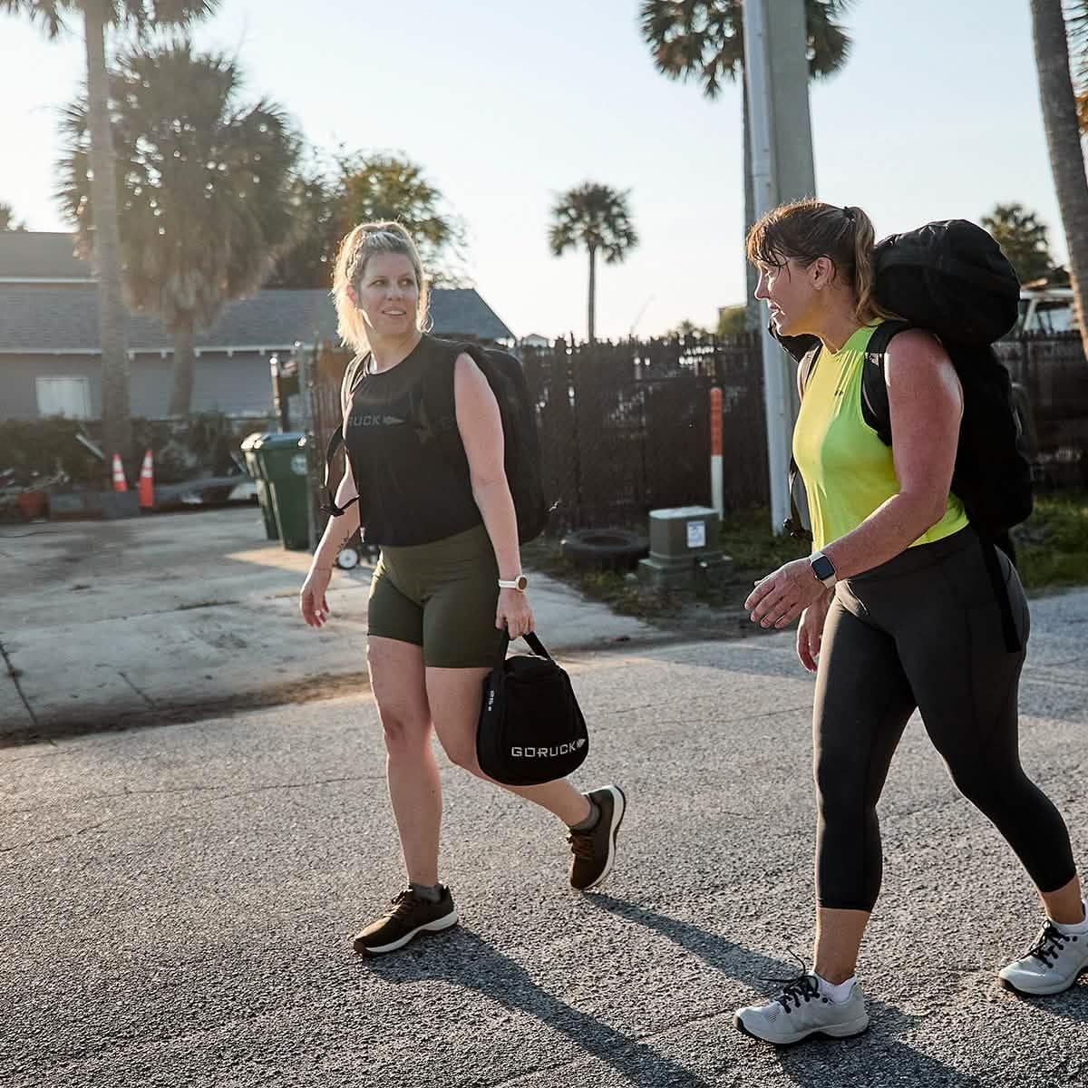 Two women rucking outdoors with GORUCK gear, wearing athletic apparel on a sunny street