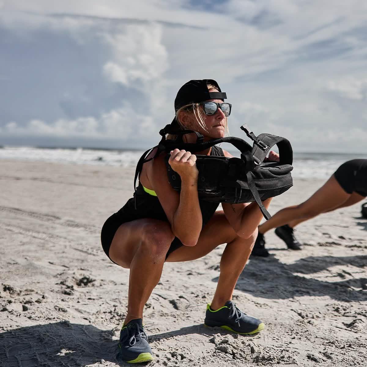 Woman training with GORUCK rucksack on sandy beach, squatting for outdoor fitness workout