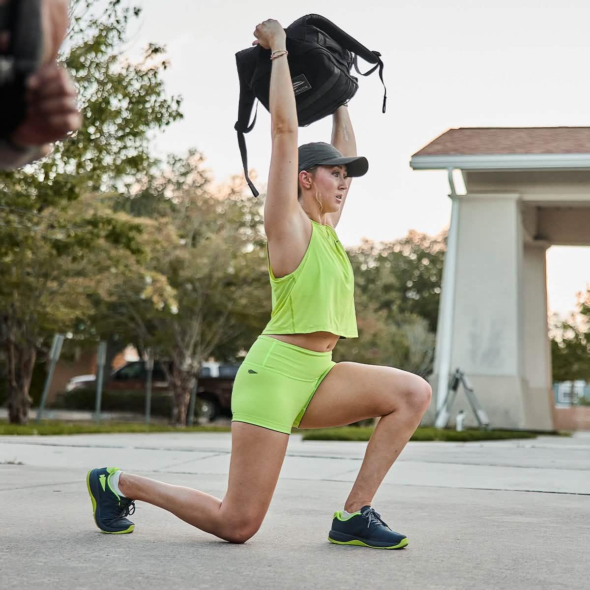 Woman in neon green workout attire lunging outdoors lifting a black GORUCK backpack overhead