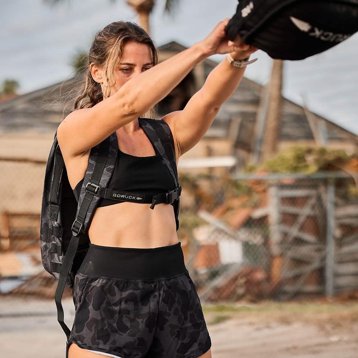 Woman wearing black GORUCK sports bra and camo black frogskin shorts lifting weighted GORUCK pack outdoors