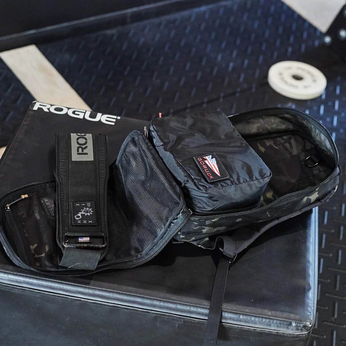 A black travel ruck made from 200D nylon rests open on a padded surface, displaying a black belt featuring a Rogue logo. Beside it, on the floor, a white weight plate is partially visible. The travel ruck includes packing cubes by GORUCK for organized storage.