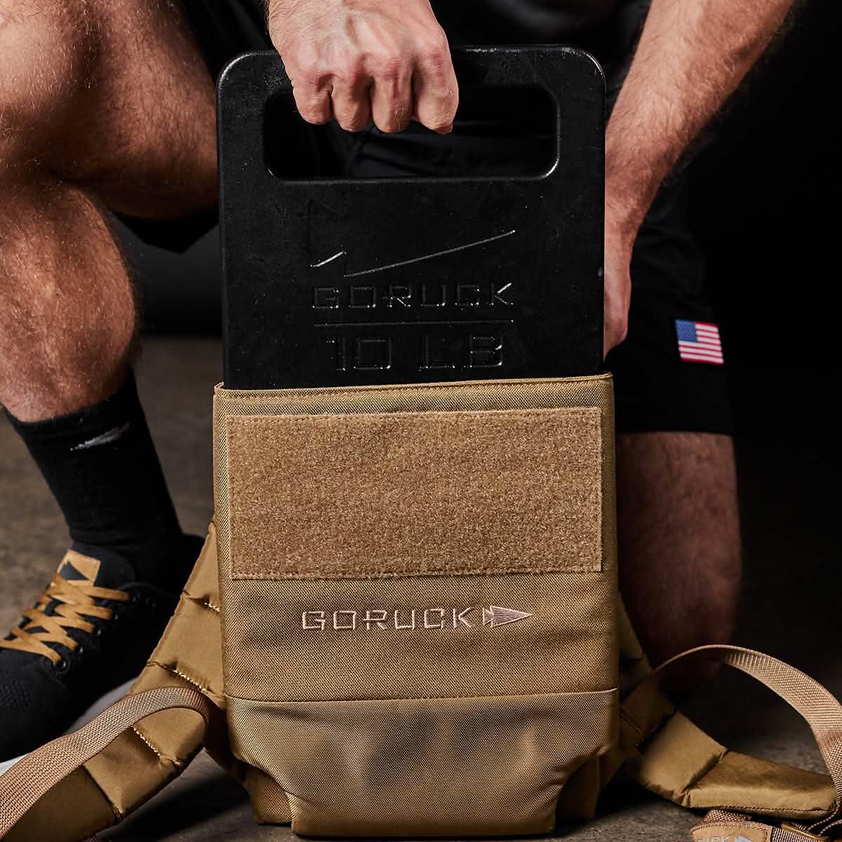A person is putting a black GORUCK 10 LB Ruck Plate into a brown GORUCK backpack. The durable Rucker backpack, equipped with Velcro and showcasing the GORUCK brand, highlights American craftsmanship. The individual is wearing black athletic shorts featuring a small American flag.