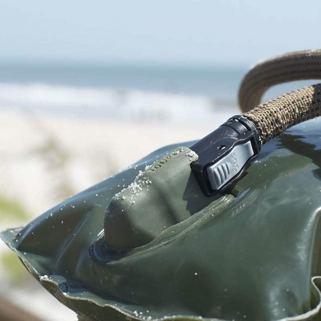 Close-up of green waterproof GORUCK bag corner with rope handle and black buckle by sandy beach