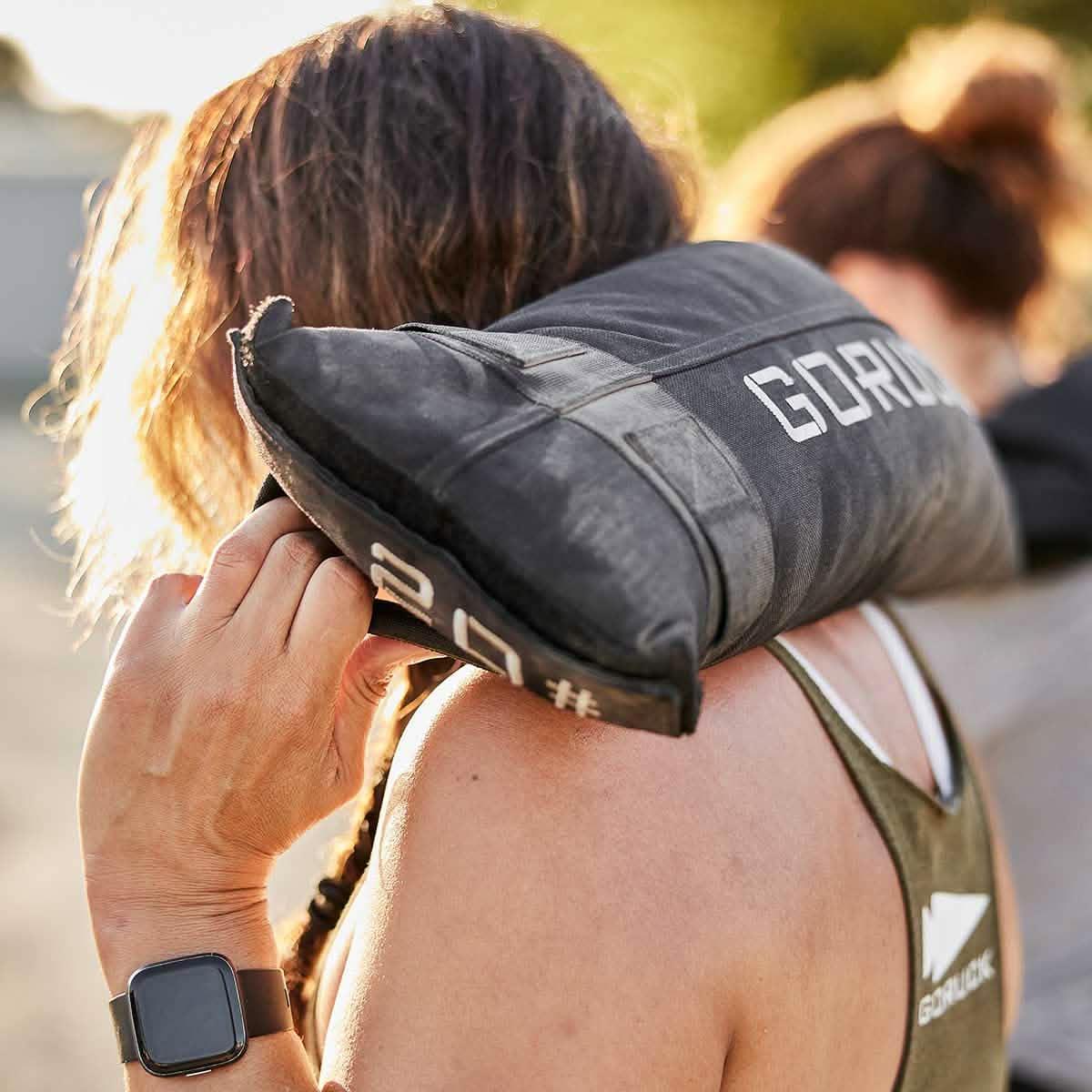 Woman outdoors carrying a GORUCK 20 lb sandbag on her shoulder, wearing GORUCK apparel.