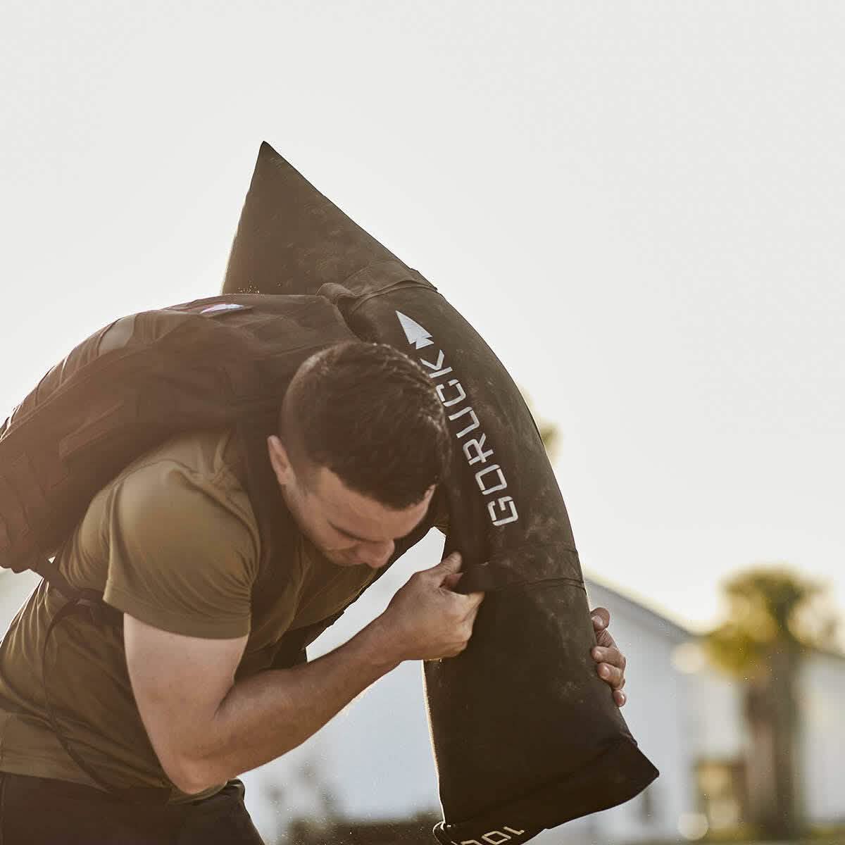 A person wearing a green shirt is outdoors, easily carrying a heavy Simple Training Sandbag from GORUCK over their shoulder using its padded handles. The blurred background suggests trees and a building beneath the bright sky, providing perfect conditions to test the durability promised by the Scars Lifetime Guarantee.