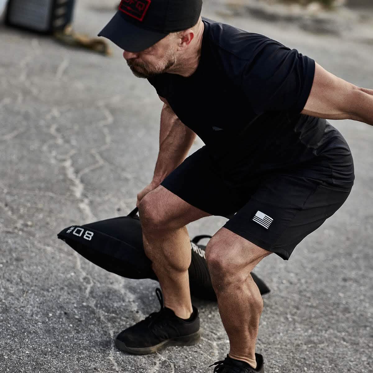 A man dressed in black athletic gear, complete with a cap, is executing a sandbag workout outside on a concrete area. Intently squatting, he skillfully manages the GORUCK Simple Training Sandbags, appreciating the padded handles that provide superior grip and comfort for high-intensity training.