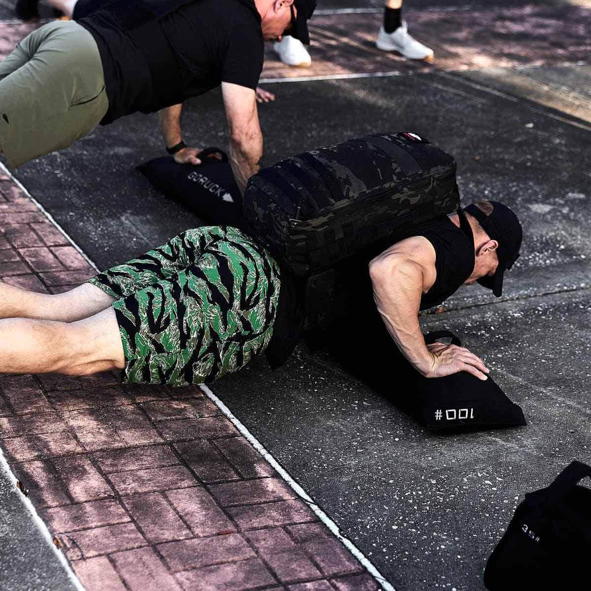 A person outdoors performs weighted push-ups with a camouflage backpack, likely filled with GORUCK's Simple Training Sandbags or gear from their home gym. Nearby, another individual mirrors the exercise. The ground is paved with bricks and asphalt. Both display dedication that reflects GORUCK's commitment to quality and persistence reminiscent of Scars Lifetime Guarantee.