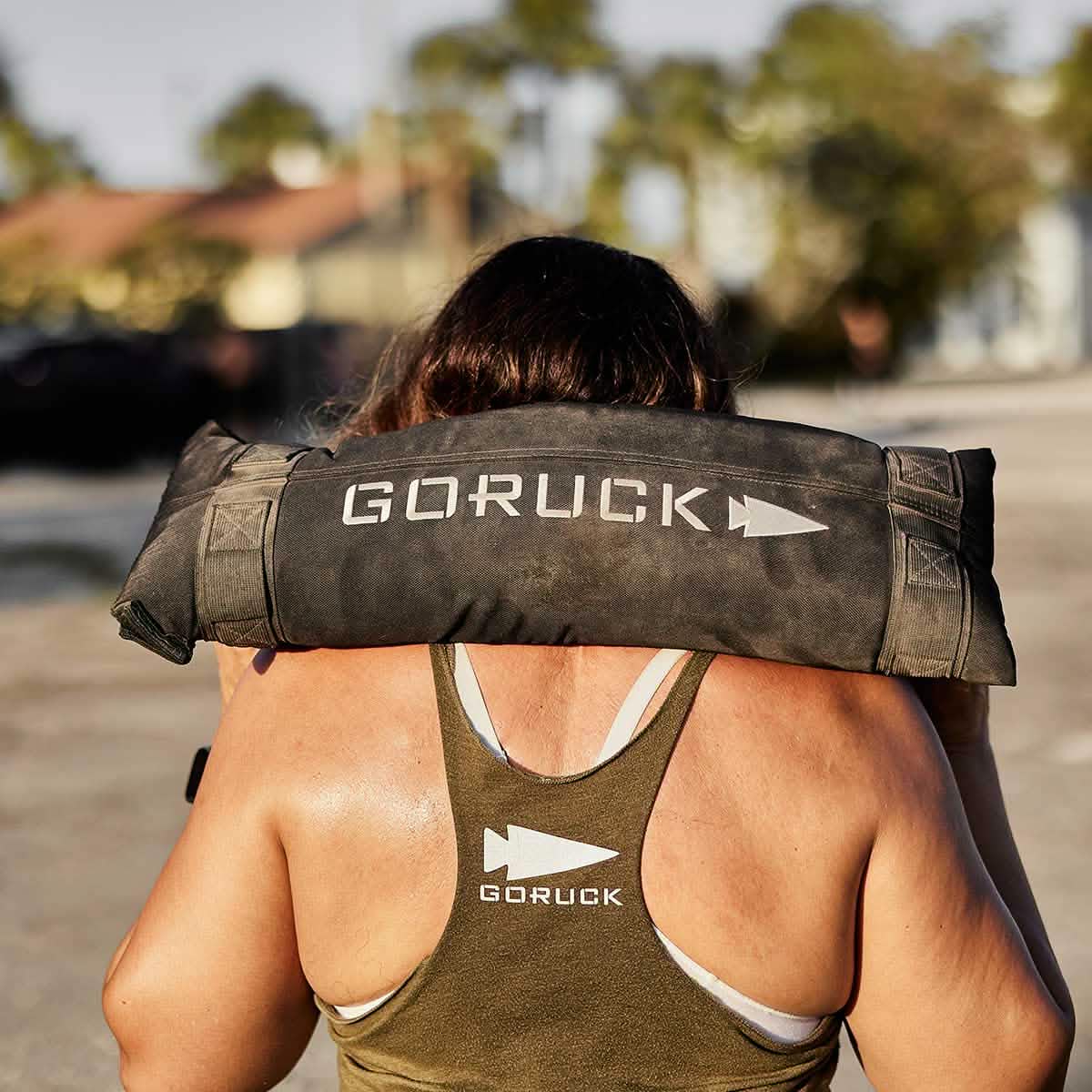 A person with long hair, wearing a green GORUCK tank top, hoists a black GORUCK Simple Training Sandbag with padded handles on their shoulders. The scene is outdoors, with blurry buildings and palm trees in the background.