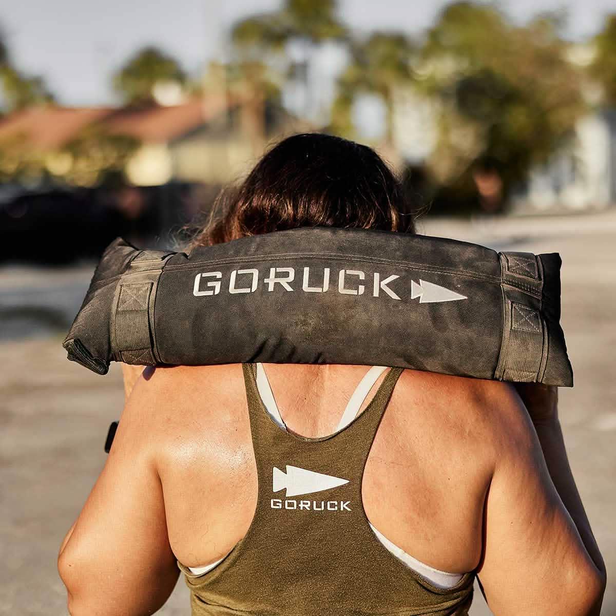 Person carrying a black GORUCK weighted sandbag on shoulders outdoors, wearing a green GORUCK tank top