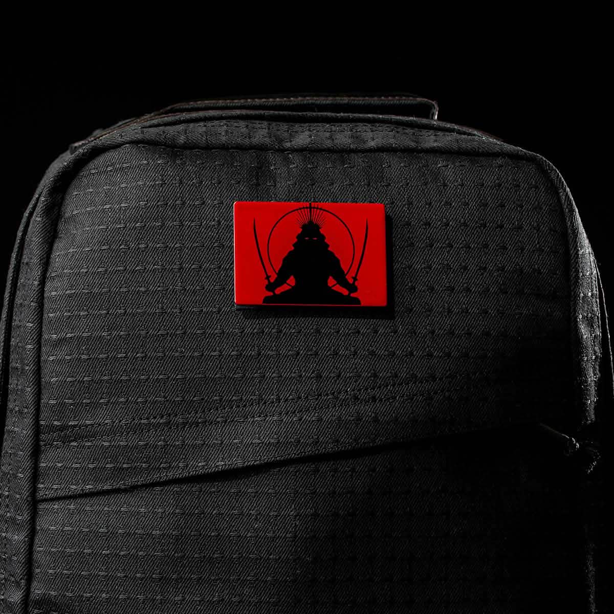 The GR1 USA x Carryology - GRXC2 Samurai is a black backpack made from durable Japanese cotton, featuring a red patch with a samurai wielding two swords.