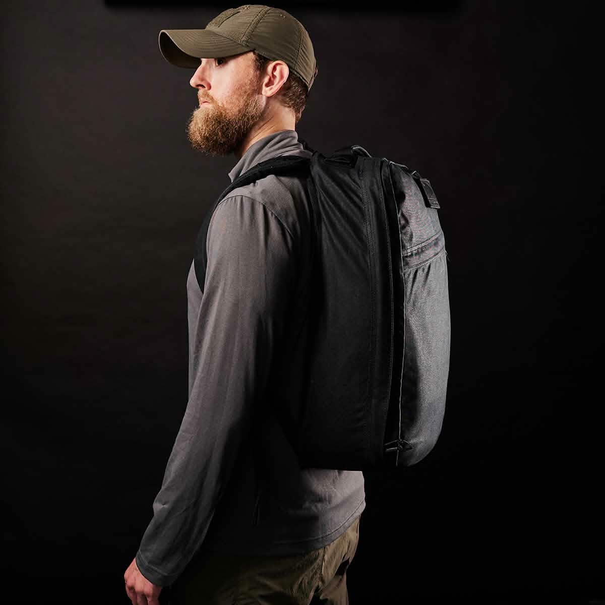 A bearded man in a cap and gray jacket is shown in profile against a dark background, showcasing the GR1 USA - Heritage by GORUCK. The large black heritage rucksack, crafted from waxed duck canvas with its Martexin Wax finish, adds a rugged touch to his look.