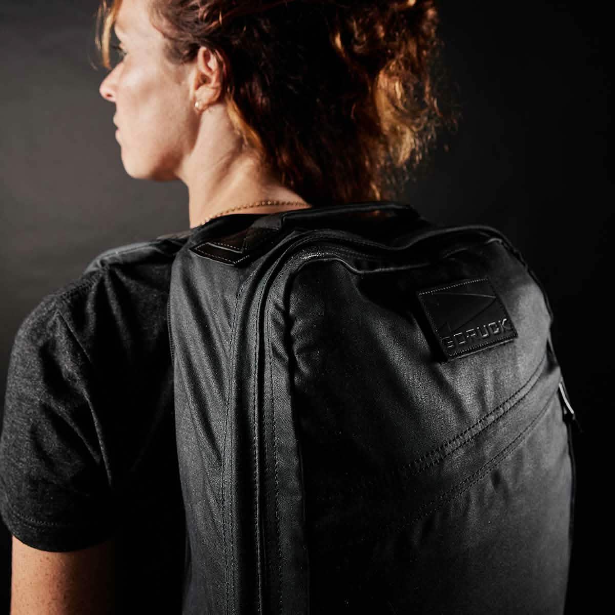 A person with curly hair is shown from the side wearing a large GR1 USA - Heritage rucksack by GORUCK. Dressed in a dark shirt, the background also remains dark, highlighting the Martexin Wax finish and texture of the backpack's design.