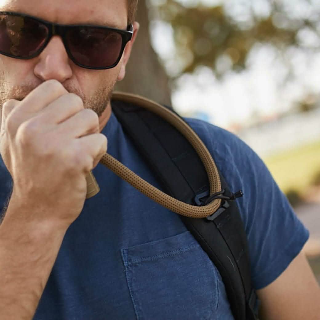 A person wearing sunglasses and a blue shirt drinks from a hydration bladder hose outdoors. The Bladder Hose Retainer by GORUCK holds the tube near their mouth, with the backpack strap visible on their shoulder. Trees and a bright sky create a backdrop that evokes the feel of GORUCK Events adventures.