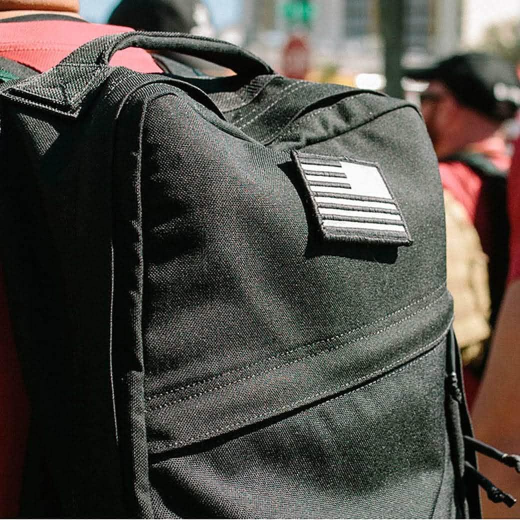 Close-up of a black GORUCK rucksack with an American flag patch, worn by a person in an outdoor urban setting