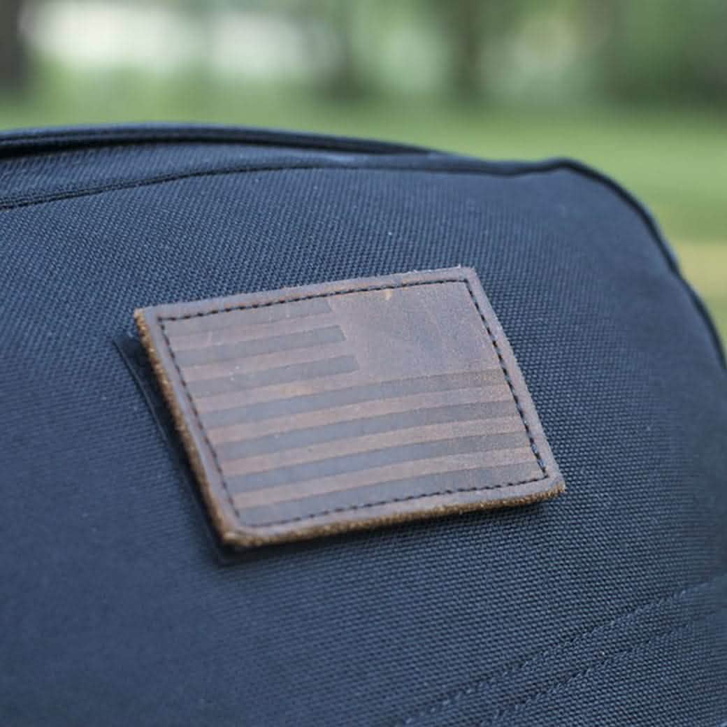 Close-up of a black fabric bag showcasing a rectangular patch with a matte, monochromatic reverse American flag design. The patch has a VELCRO® backing and is from the brand vendor-unknown. The background features blurred green foliage.