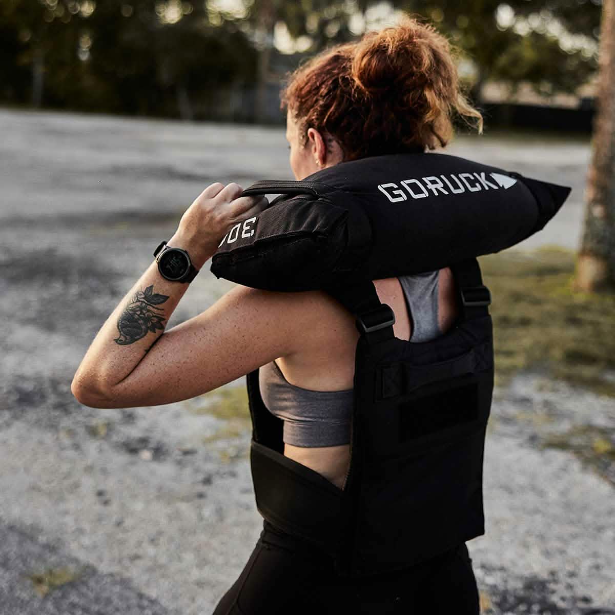 A person with a tattoo on their left arm carries a Simple Training Sandbag by GORUCK over their shoulders. They are wearing a fitness vest and standing outdoors on a gravel surface with trees in the background, embodying the rugged spirit of the Scars Lifetime Guarantee.