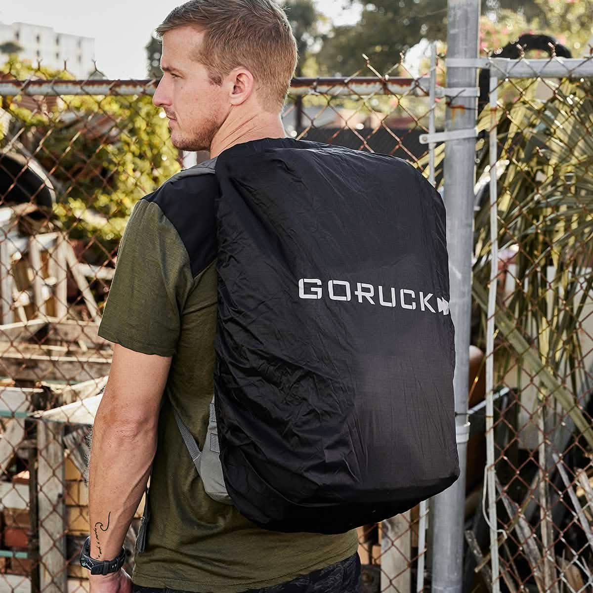 A man is outdoors wearing a green shirt and carrying a large black GORUCK backpack, which is protected by the Ruck Rain Cover. Behind him, a chain-link fence subtly frames the scene with some foliage in the background.