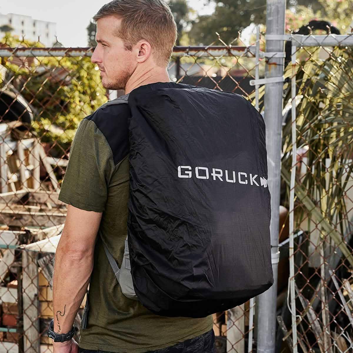 Man wearing black Goruck backpack cover outdoors near chain-link fence with greenery