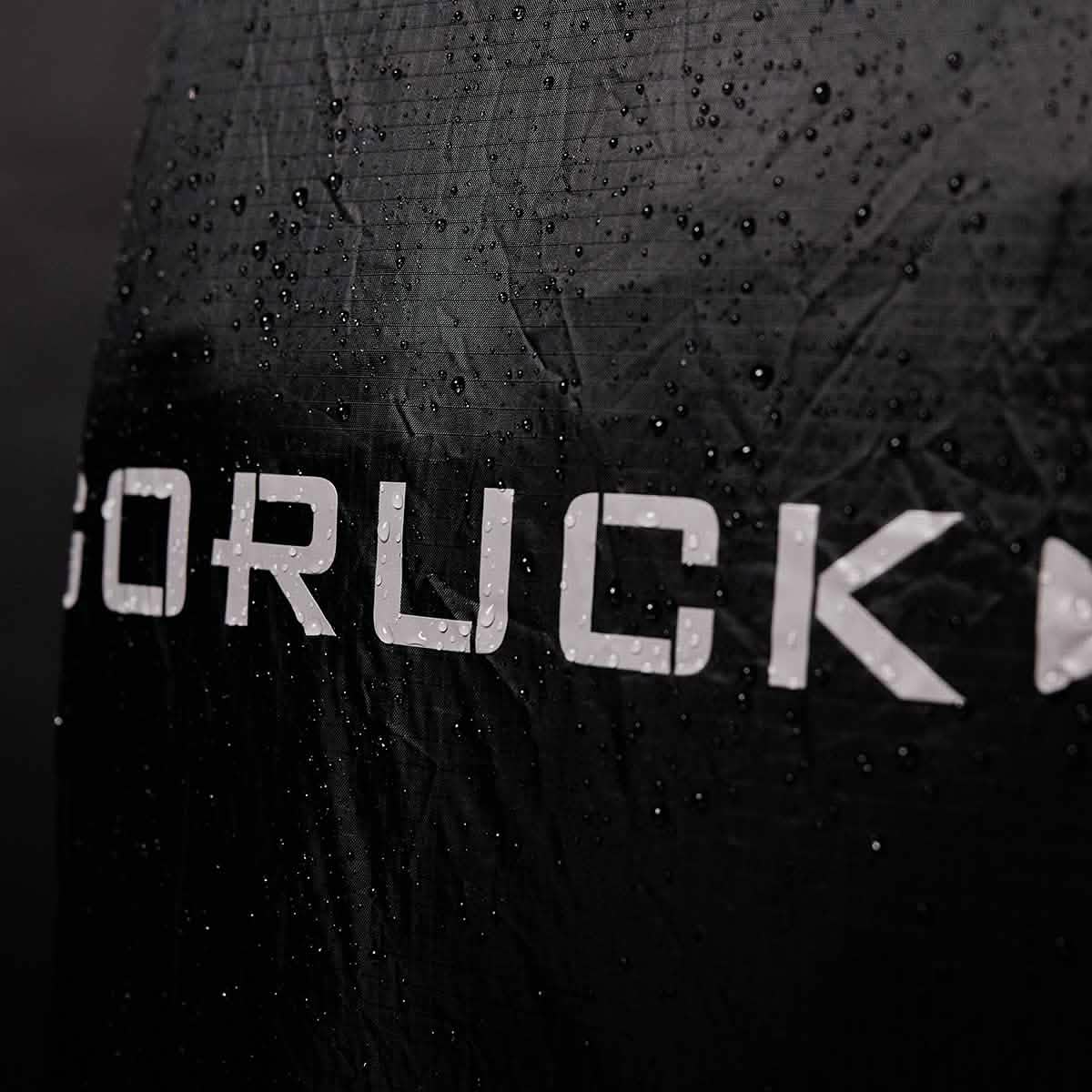 Close-up of a black fabric featuring the GORUCK logo prominently in bold white letters, with water droplets scattered across its surface. This suggests the lightweight, waterproof design of the Ruck Rain Cover, creating a textured appearance against a slightly blurred background.