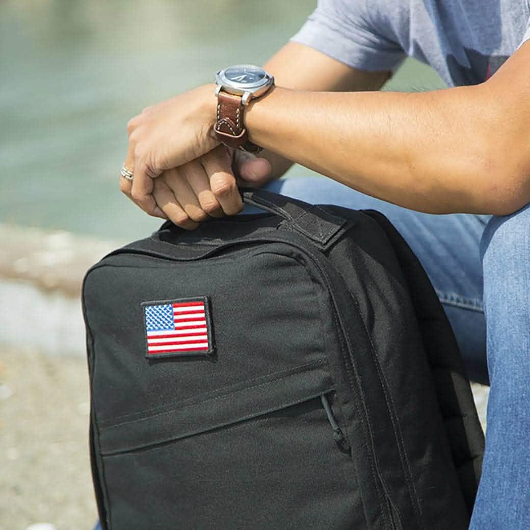 Person with watch holding black rucksack featuring a US flag patch, outdoors near water