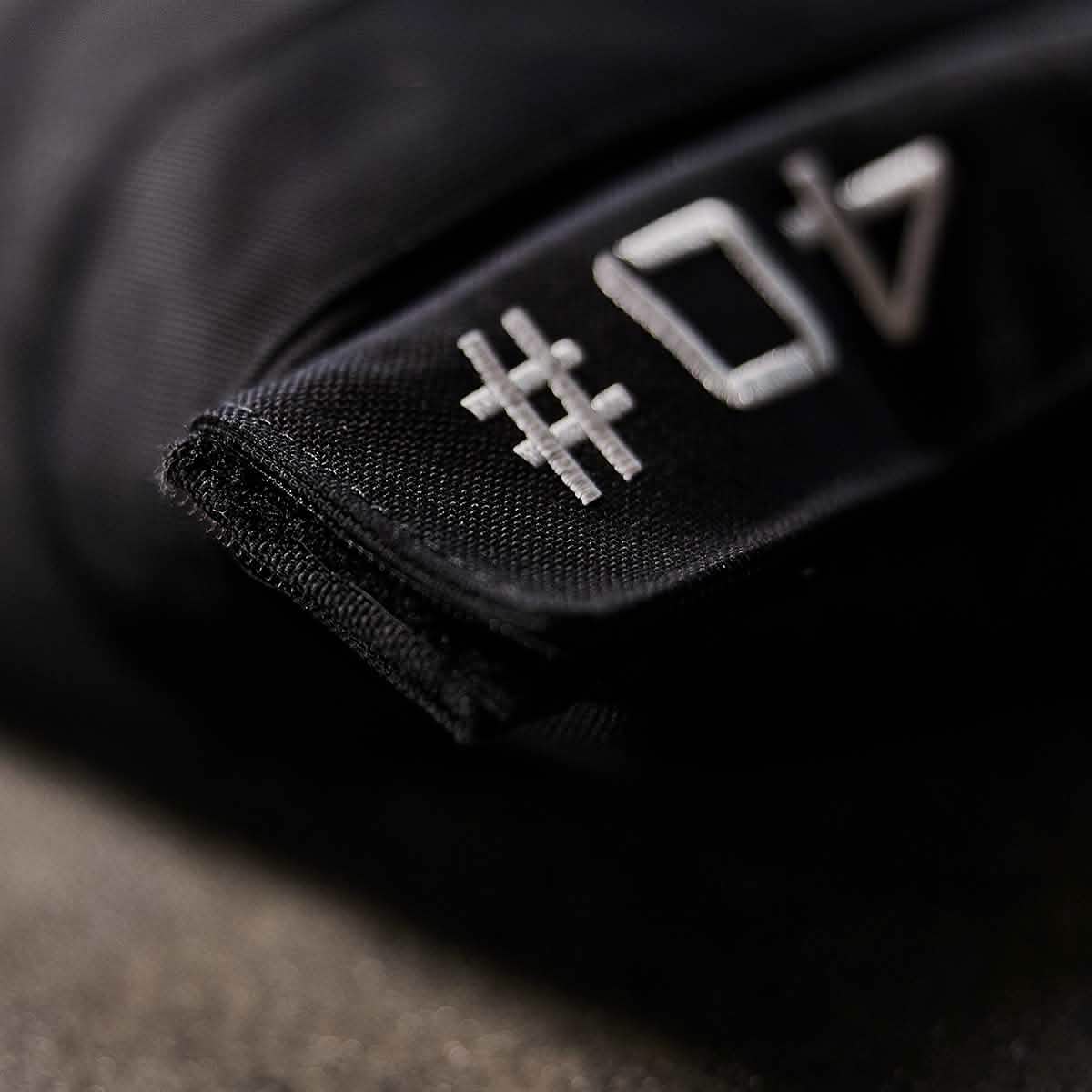 The image features a close-up of GORUCK's Sandbags 2.0 in black fabric, showcasing white lettering with the characters #04. Made from tough 1000D CORDURA, the fabric has a rugged texture and prominently embroidered numbers, indicating robust construction typical of durable clothing or accessories. The softly focused background adds an aesthetic touch.