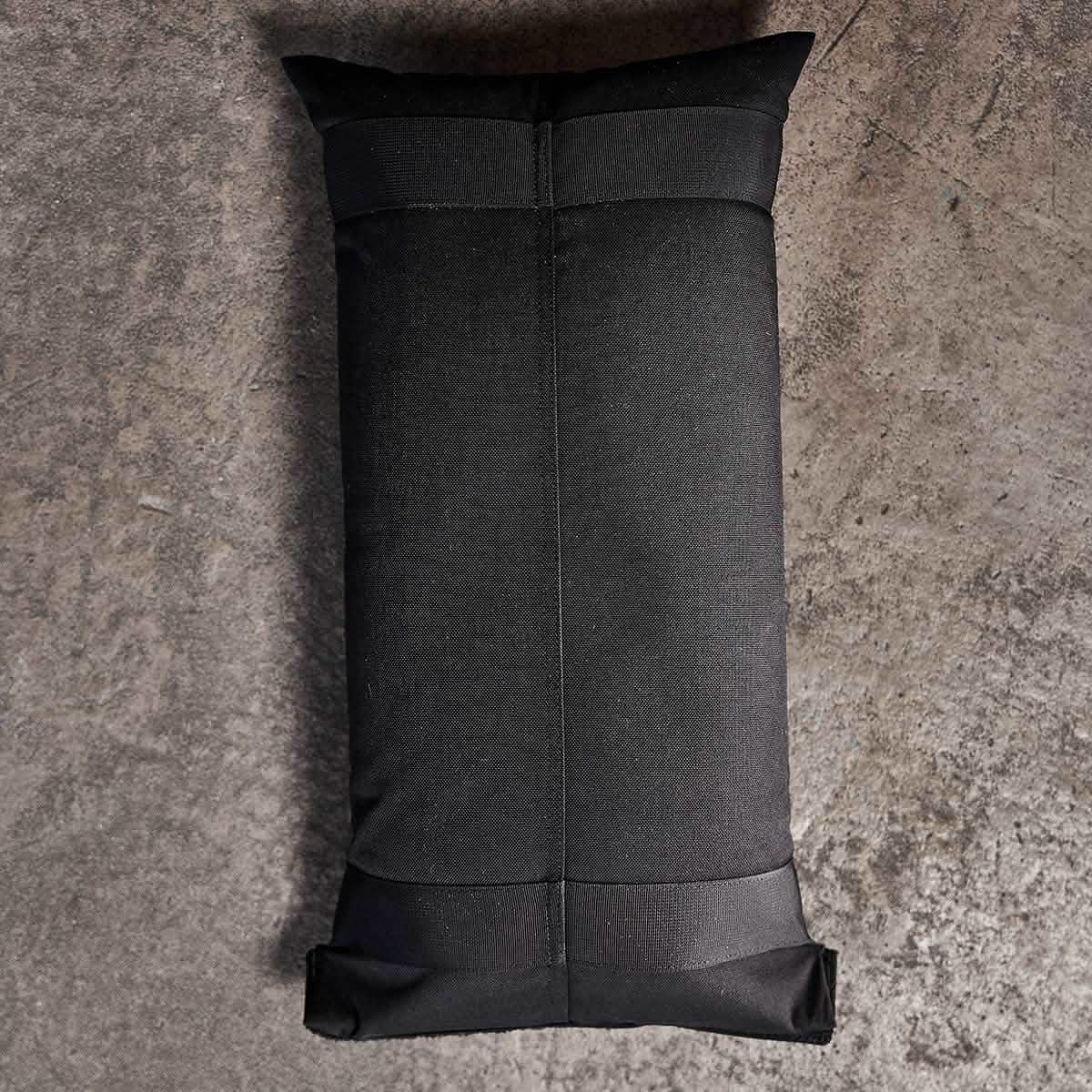 The GORUCK Sandbags 2.0, with their simple and sturdy design, lay on a rough-textured gray concrete floor in a top-down view. This rectangular black weighted bag, made from rugged 1000D CORDURA, showcases visible seams and reinforced corners, making it ideal for training sandbags.