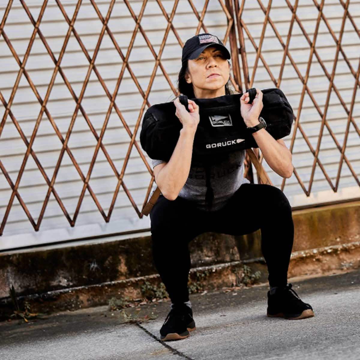 Dressed in athletic wear and a cap, an individual squats with a weighted GORUCK Sandbags 2.0 backpack, celebrated for its 1000D CORDURA durability. They are exercising outdoors on a concrete surface, with a diamond-patterned metal fence as the backdrop.