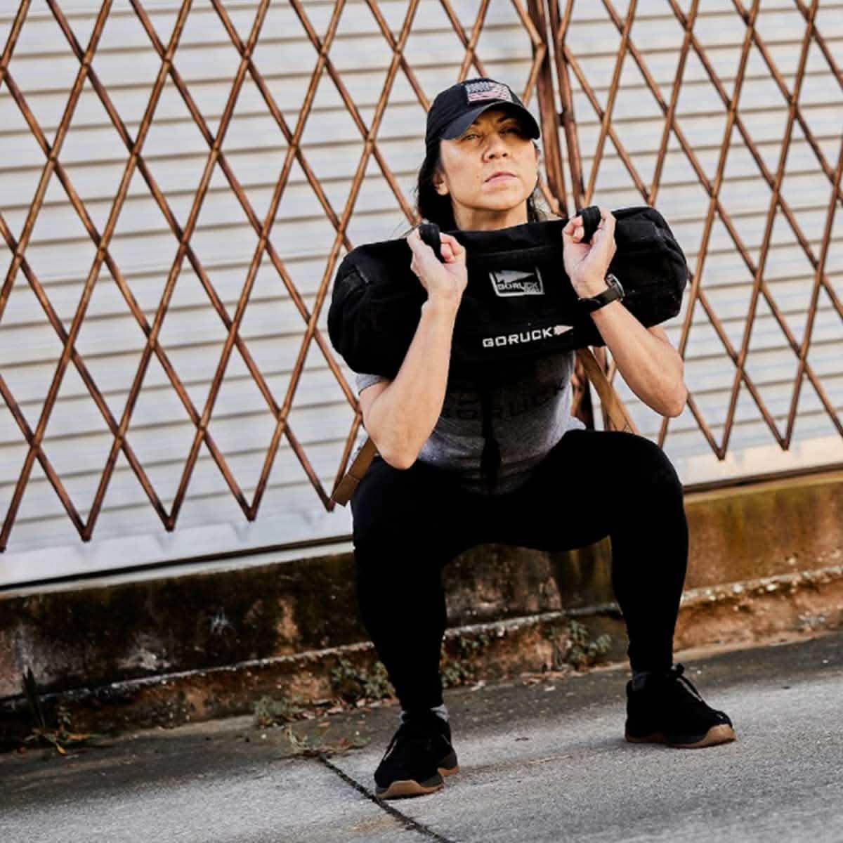 Woman squatting outdoors with GORUCK sandbag, wearing athletic gear and a GORUCK cap.