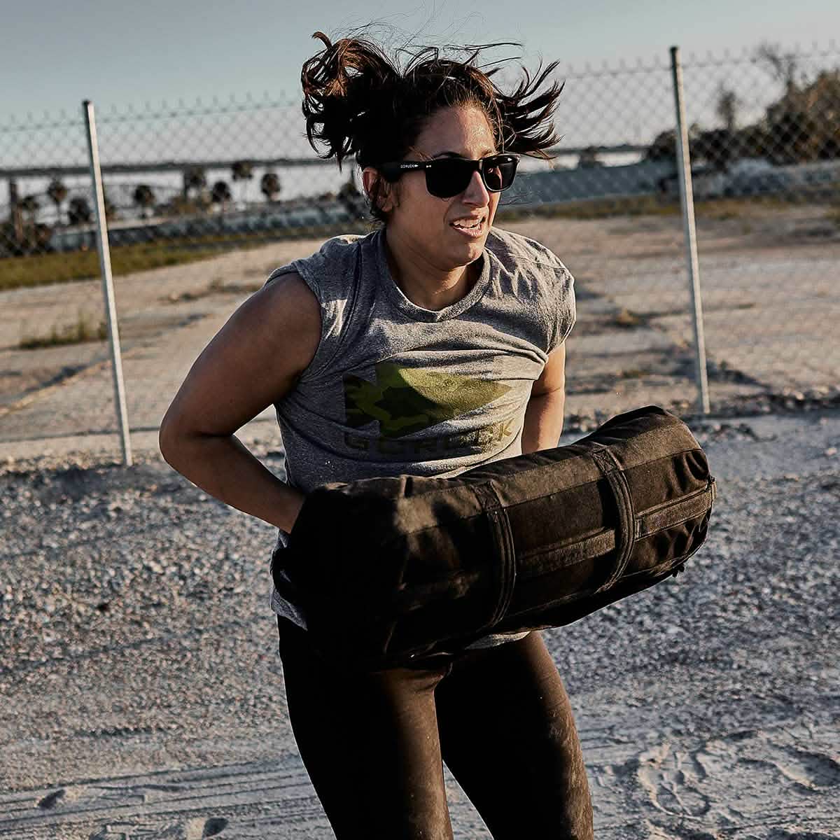 Wearing sunglasses and a gray sleeveless shirt, a person exercises outdoors with the Sandbags 2.0 from GORUCK, made from durable 1000D CORDURA. They are on a dirt path with a chain-link fence in the background. Their tousled hair suggests movement and intensity.