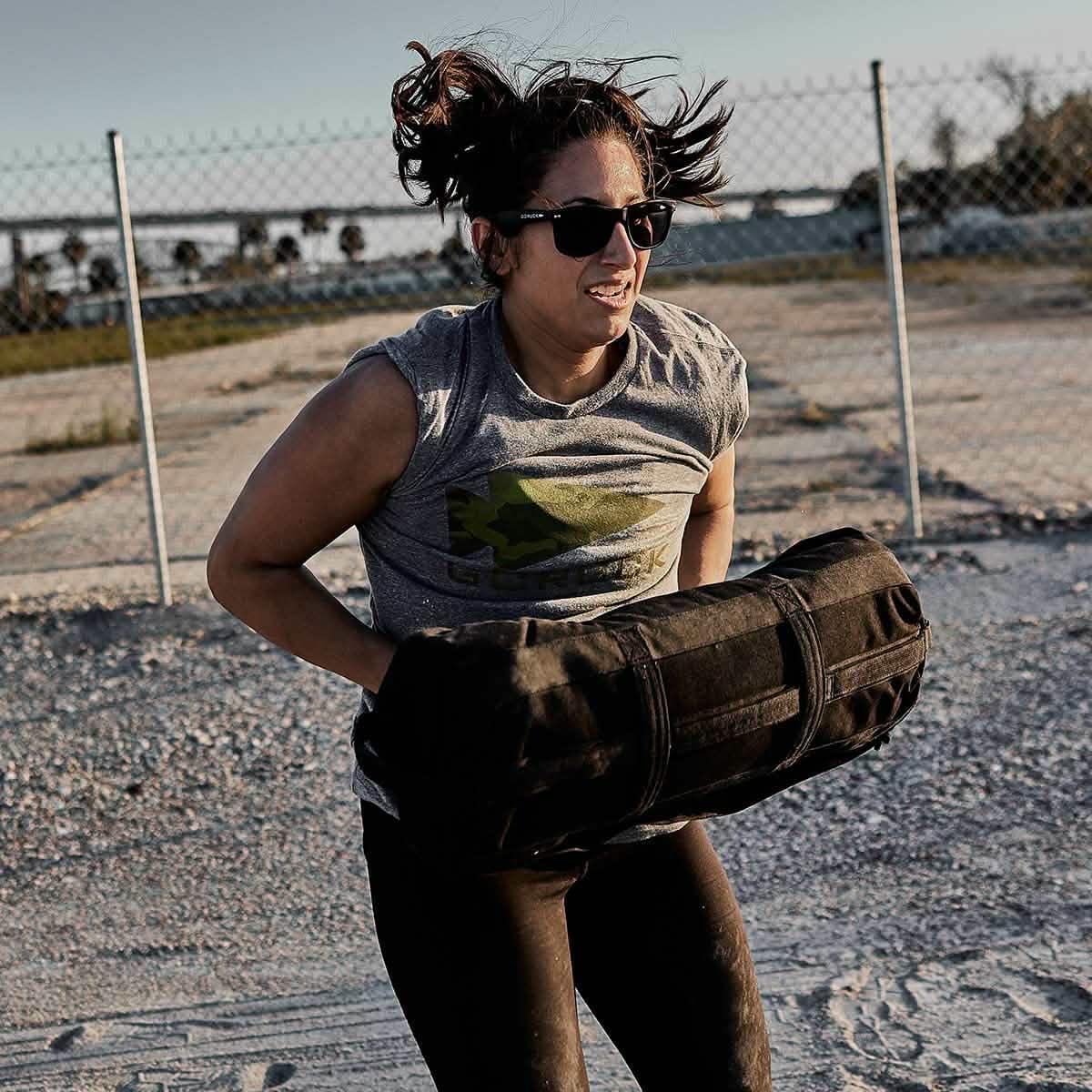 Woman wearing sunglasses and GORUCK shirt lifting a heavy black rucksack outdoors on gravel