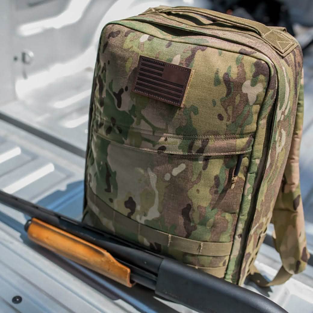 A truck bed holds a camouflage backpack featuring a vendor-unknown Patch - Leather Reverse Flag attached with VELCRO®, while a shotgun with a wooden handle lies beside it.