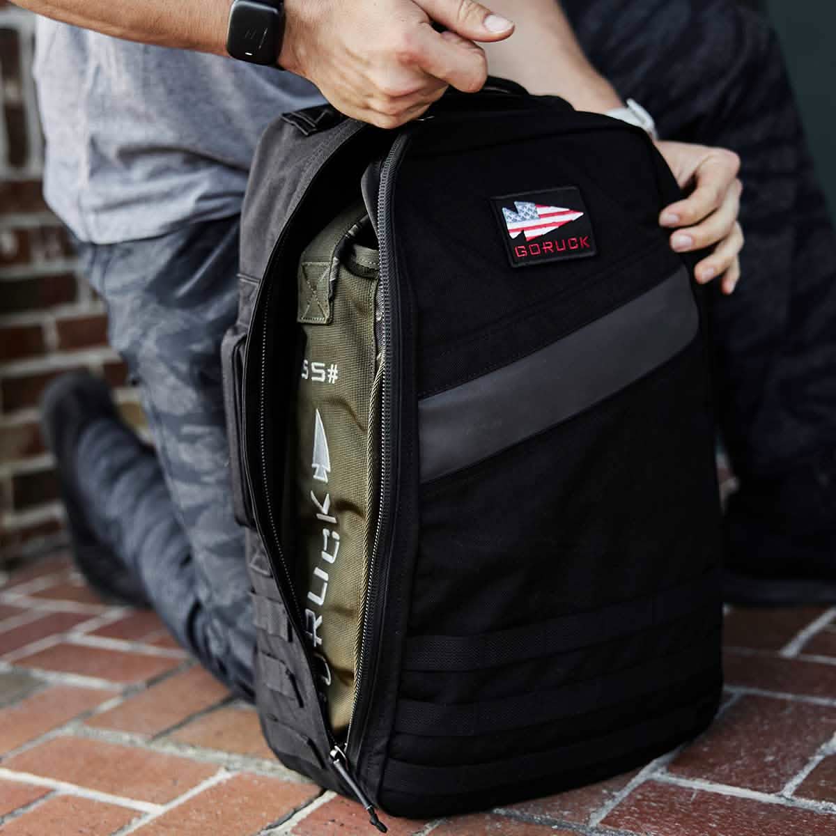 A person kneels on a brick surface, unzipping a black GORUCK backpack made from Special Forces Grade materials, revealing a Sand Jerry Can inside. The backpack is adorned with an American flag patch and black MOLLE webbing. The person's wristwatch is visible.