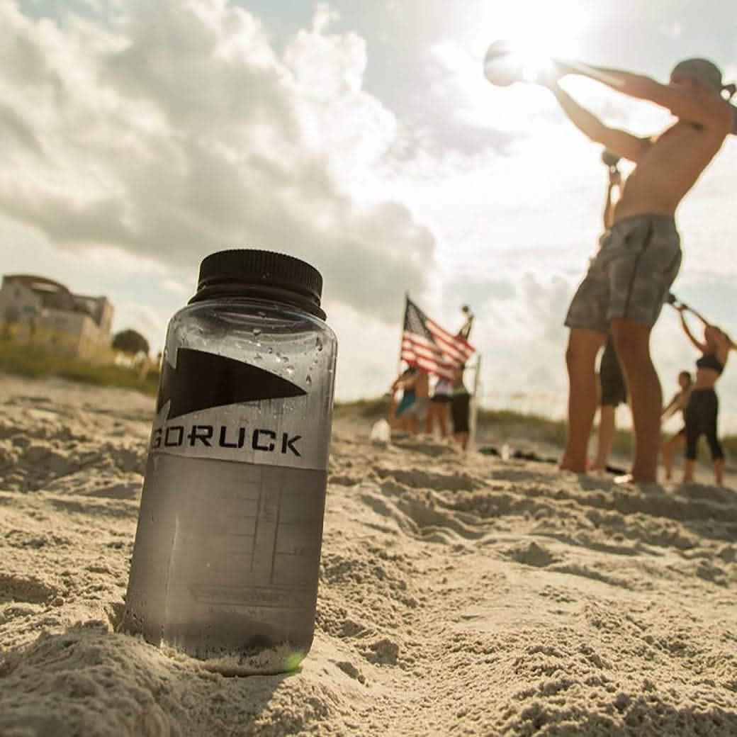 GORUCK water bottle in sand with fitness group exercising and American flag in background