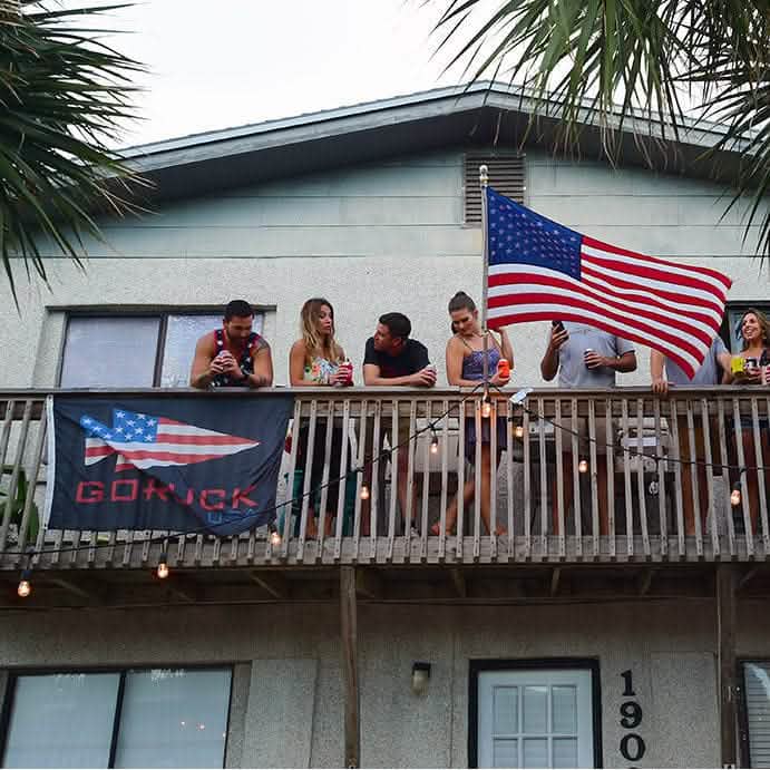 A group of people stands on a balcony decorated with weather-resistant nylon GORUCK USA flags by Annin Flagmakers, enjoying a casual gathering beneath the glow of string lights.