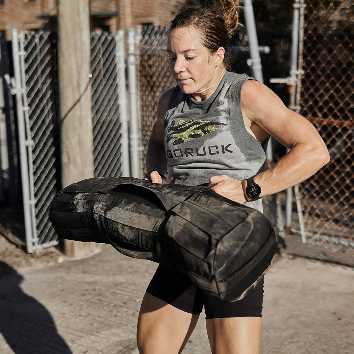 A woman in a sleeveless top and shorts works out outdoors, training with Sandbags 2.0 by GORUCK, crafted from durable 1000D CORDURA. She is focused and appears to be exerting effort. The background features a chain-link fence and a wooden pole, embodying the intensity of her routine.