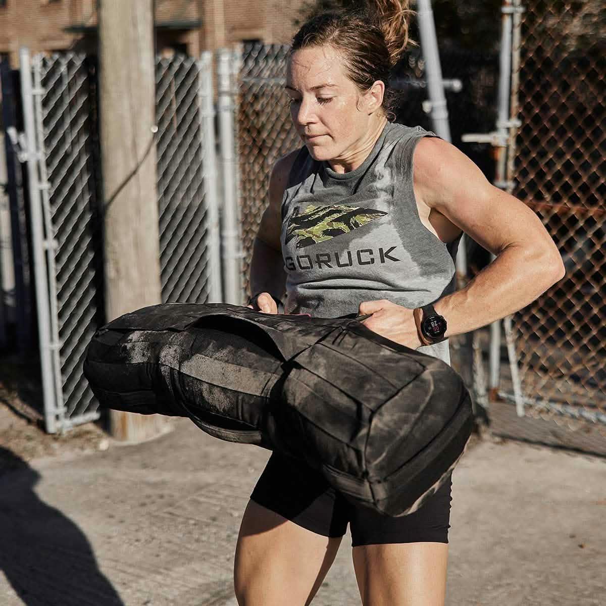 Fit woman lifting heavy black 80lb GORUCK sandbag outside near chain-link fence during intense workout