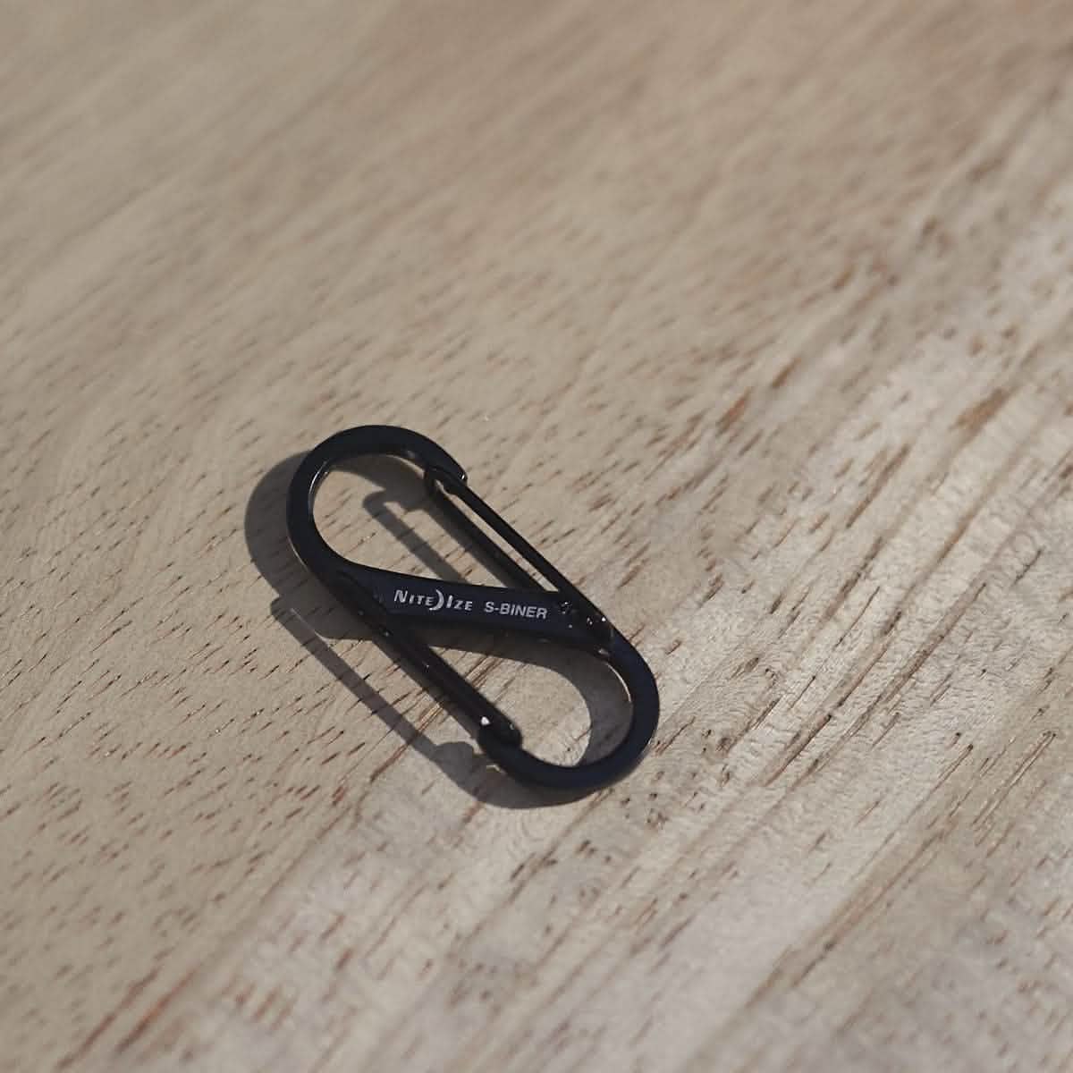 A compact black metal S-shaped carabiner sits on a wooden surface, displaying the Nite Ize brand. Ideal for attaching to your rucksack, this Carabiner - Hydration Bladder Attachment features a spring-loaded gate on each end, designed to connect items securely.