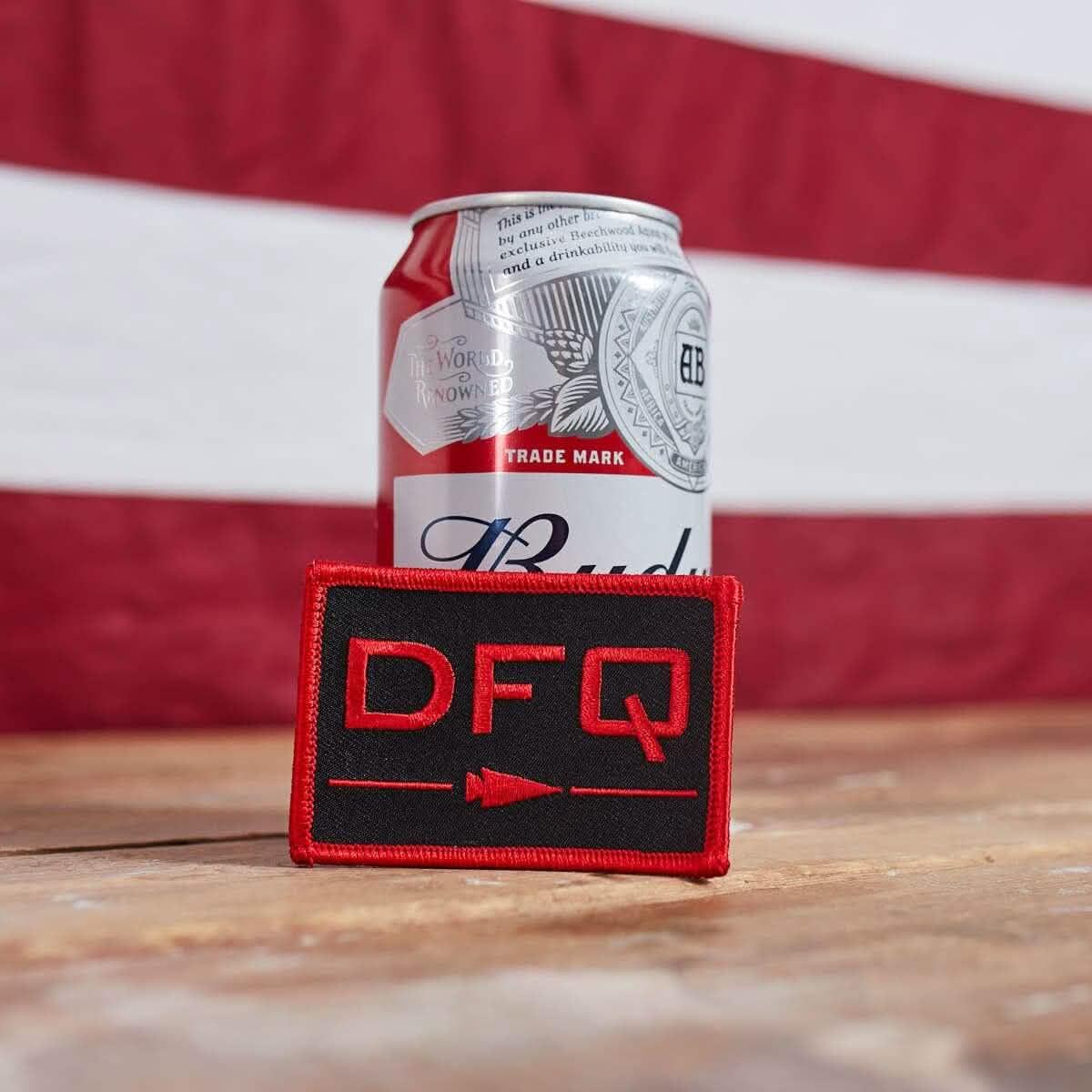 Red and black DFQ patch displayed in front of a Budweiser beer can on a wooden surface with red and white background