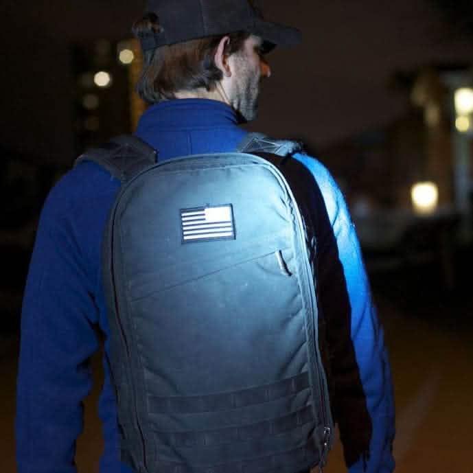 A man wearing a cap and a blue sweater strolls at night with his black backpack, adorned with the Reflective Reverse Flag Patch from vendor-unknown. The patch's reflective material catches the light from blurry building lights in the background.