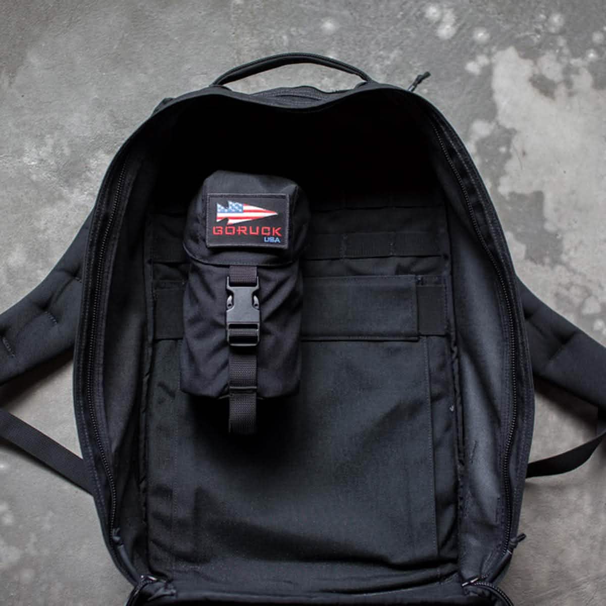A black backpack from GORUCK with an open main compartment rests on a concrete floor, highlighting its MOLLE attachment system. Inside, a smaller black pouch is secured, displaying an American flag patch with the words